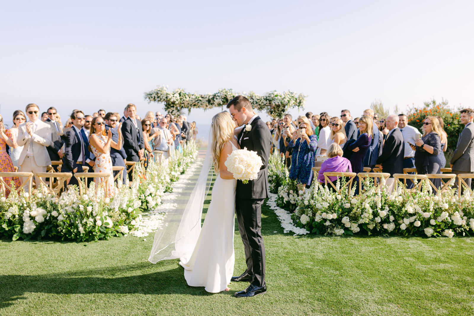 A kiss in the aisle as the guests stand, white florals down the lawn at Terranea